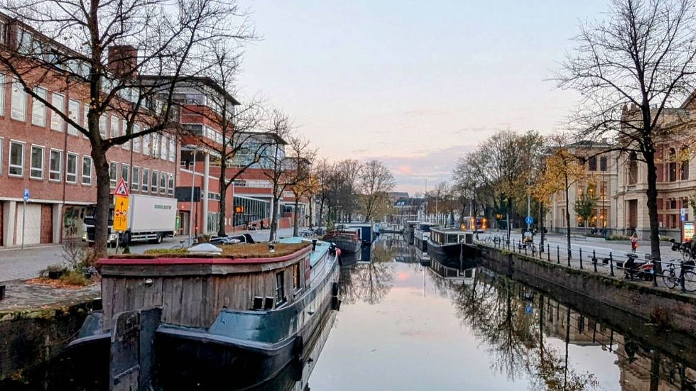 Canal with moored boats reflects trees and city buildings at dawn. A truck passes on a street lined with bare trees and bicycles.