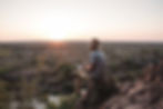Man in a hat sits on a rocky hill at sunset on Safari, holding a tablet, overlooking a vast green landscape. Warm, tranquil ambiance.