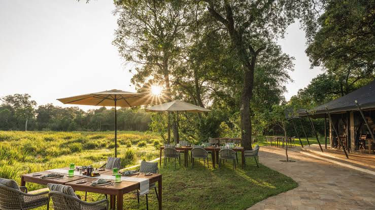 Outdoor dining area at a Wilderness safari camp surrounded by mature trees and open grassland.
