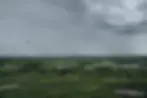 A helicopter flies over a vast, green landscape with scattered trees and water patches under a cloudy, gray sky, conveying a moody atmosphere.