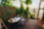 Woman in a bubble bath holding a drink on a wooden deck, surrounded by lush greenery at sunset. Man stands in the background. Relaxed mood.