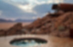 Desert Whisperer landscape with a circular pool on a wooden deck. A modern cabin sits on rocky terrain. Cloudy sky with sunlight breaking through.