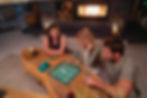 Three people play Scrabble at a wooden table near a lit fireplace. Candles and wine glasses set a cozy ambiance. Green tiles are visible.