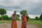 Kenyan Masai Warriors in traditional outfits in colorful robes smiling and posing outdoors on a dirt path. Lush greenery and a cloudy sky form the background. On Safari in Kenya Masai Mara