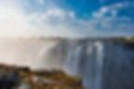 Waterfall cascading over cliffs with mist rising. Victoria falls Blue sky with scattered clouds above, rocky foreground, and distant trees. Serene atmosphere.