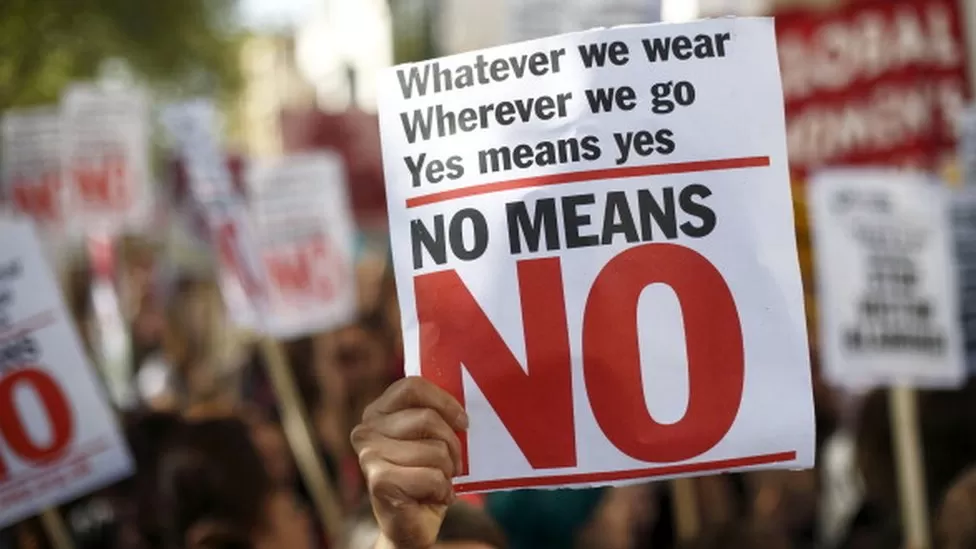 A protest sign being held up, saying "Whatever we wear, Whereever we go, Yes means yes, No means No"