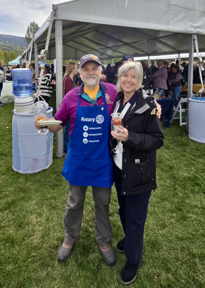 Gus and Melanie smiling behind a table of wine bottles at the Snowmass Wine Festival, with blurred attendees mingling in the background.