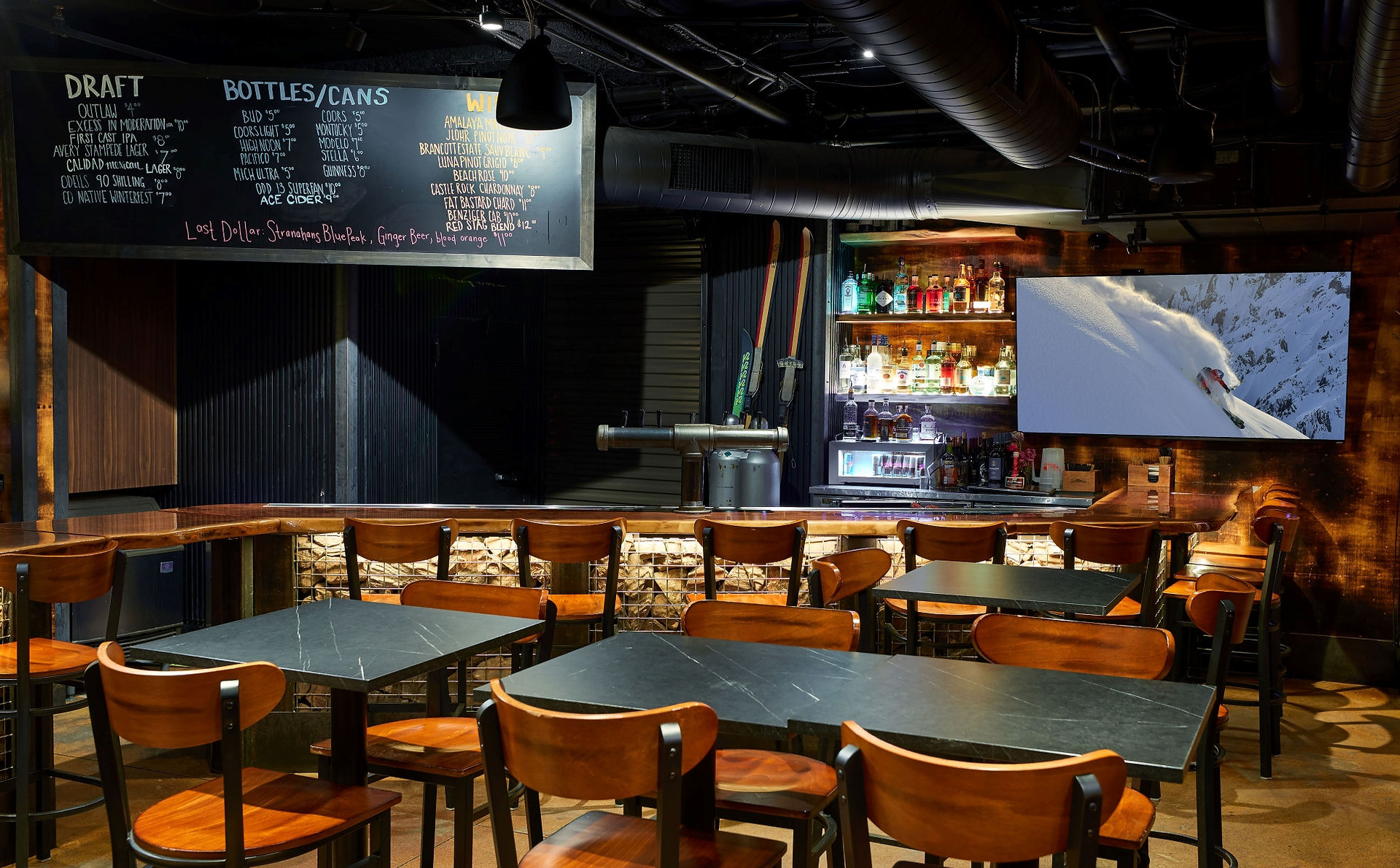 Main bar area at Buck Bar & Kitchen, showing the drinks menu board, stone gabion wall base, vintage wood finishes, and a large screen featuring skier imagery.