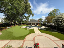 Large backyard synthetic turf putting green with multiple holes, landscaped beds, pool, and patio
