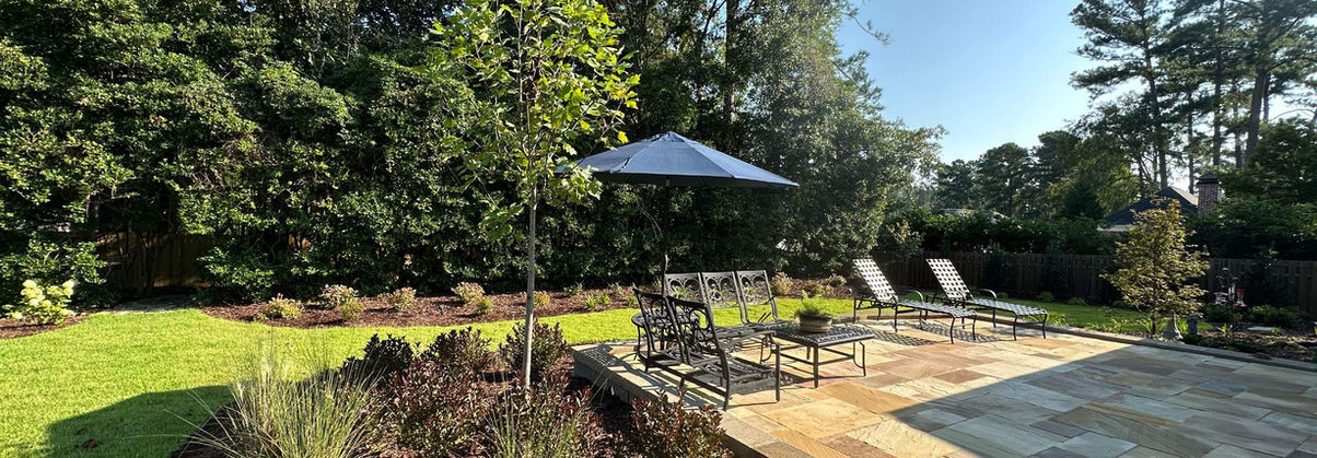 Natural stone backyard patio with lounge chairs, umbrella, and mulched planting beds; residential installation in Augusta, GA