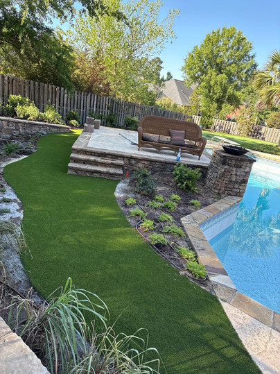 Backyard with pool decking, synthetic turf, a curved flower bed, and a partially visible swimming pool