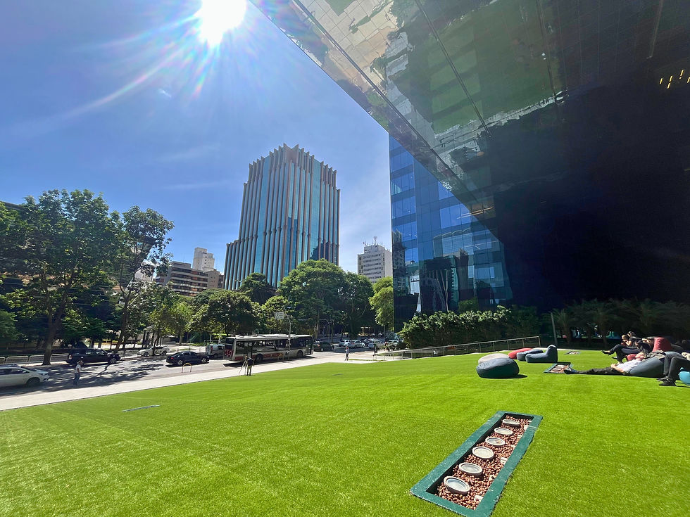 Área externa do escritório na Faria Lima, São Paulo com gramado verde, pessoas relaxando em pufes. Edifício moderno ao fundo, céu claro e sol forte iluminando a cena.