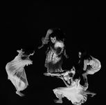 Four women in white dresses dance in unison in a circle around a worried woman in a white dress.