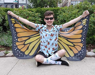 A man with short brown hair and black sunglasses sits on the sidewalk in front of some bushes. He is wearing a blue button-up shirt with monarch butterflies on it, black shorts, white socks with trans flag butterflies, black shoes, a necklace, a rainbow bracelet on his right wrist, and a watch on his left wrist. His arms are spread to show off a pair of large monarch butterfly wings of his own.