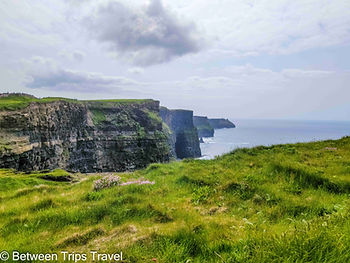 The Cliffs of Moher drop their full 700 feet into the Atlantic in a single unbroken line, the grass at the edge so green and the sky so unsettled that the boundary between land and weather feels genuinely negotiable. Photo by Between Trips Travel.