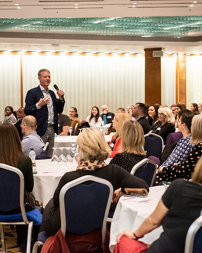Jon speaking to a large audience who are seated around tables in a conference room