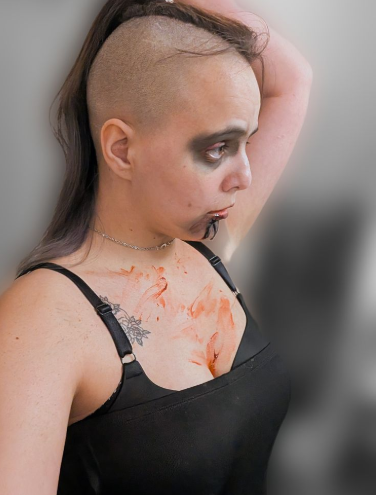 A profile view of a woman with a heavily blurred background. Woman's side of head is shaved though a high ponytail can be seen at the top of the head. Woman has on heavy dark grey/black eyeshadow in the smokey eye style with bright magenta sparkly makeup on the eyelids. There is a serious expression. Woman has blood dripping from lips. The chest is covered with blood smears and a black push up bra and black sleeveless dress that stops at where the black bra strap meets the dress. the skin tone is light olive. There is a black outline tattoo of a skull and flowers under the right bra strap on the collar bone.