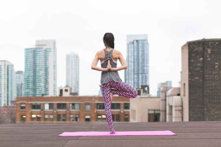 A woman standing on one leg on a pink yoga mat, on a roof of a building with a skyline in the background as she meditates.