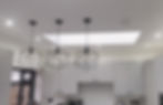 A kitchen with a black and white colour scheme with 3 black pendant lights neatly arranged over the island as well as downlights producing daylight coloured light, The image also shows a wired interlinked heat detector.