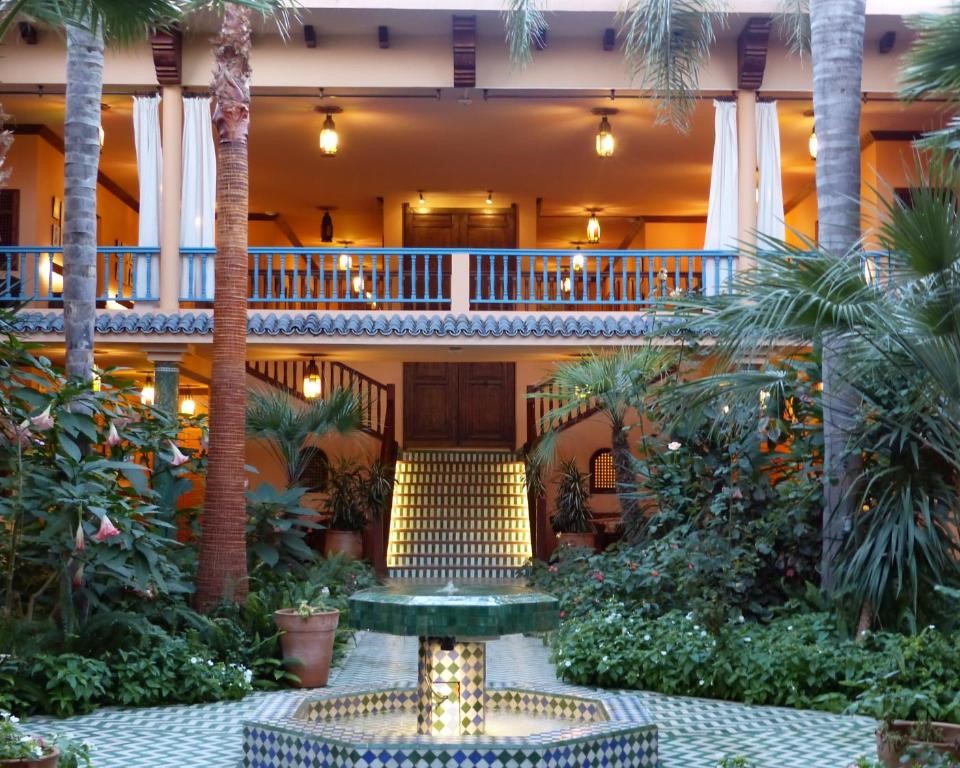 Courtyard with green and blue tiled fountain, surrounded by lush plants, palm trees, and a two-story building with warm lighting and balconies.