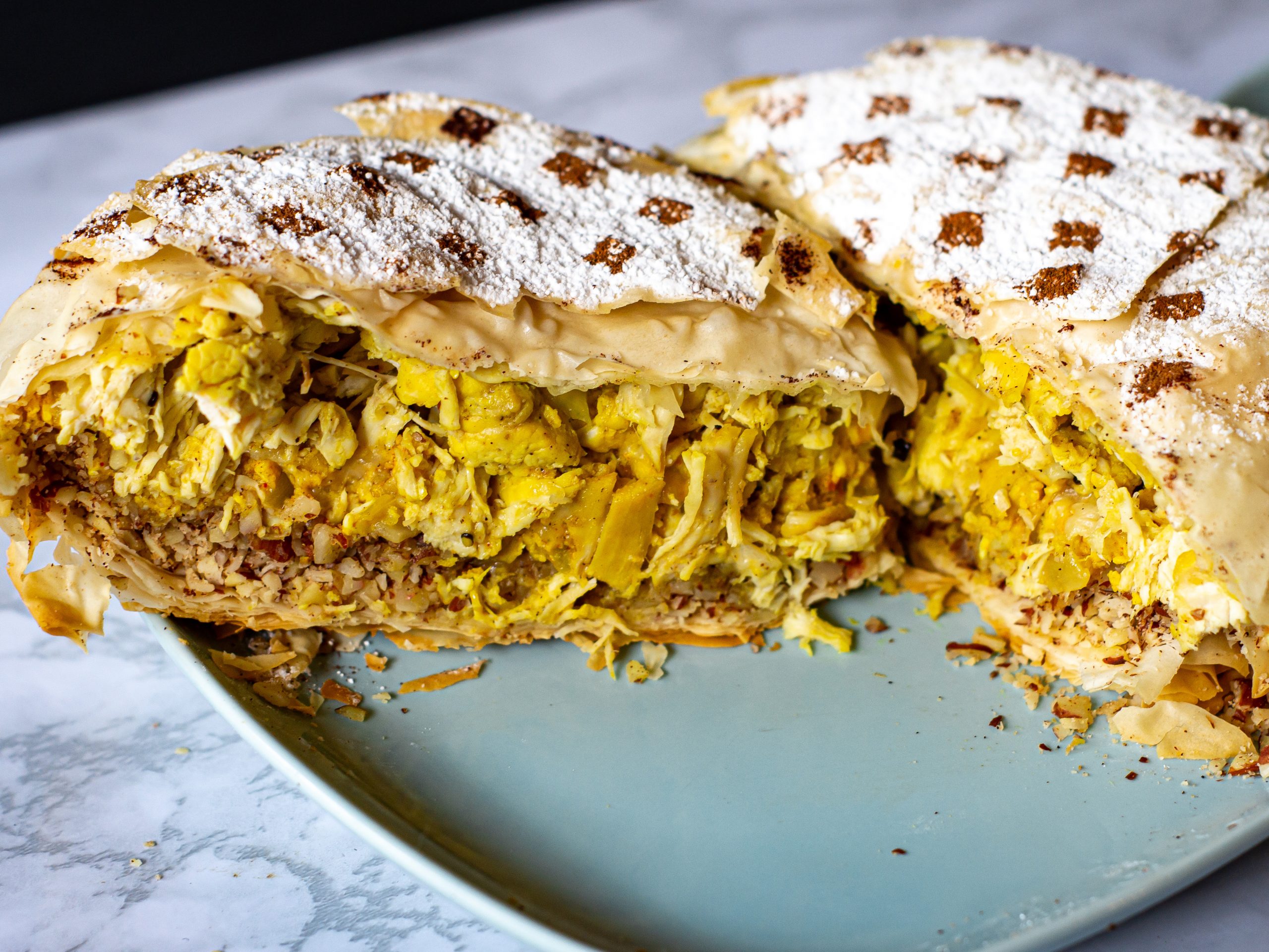 A sliced Moroccan pastilla on a light blue plate, filled with yellow chicken and nuts, topped with powdered sugar and cinnamon, on marble.