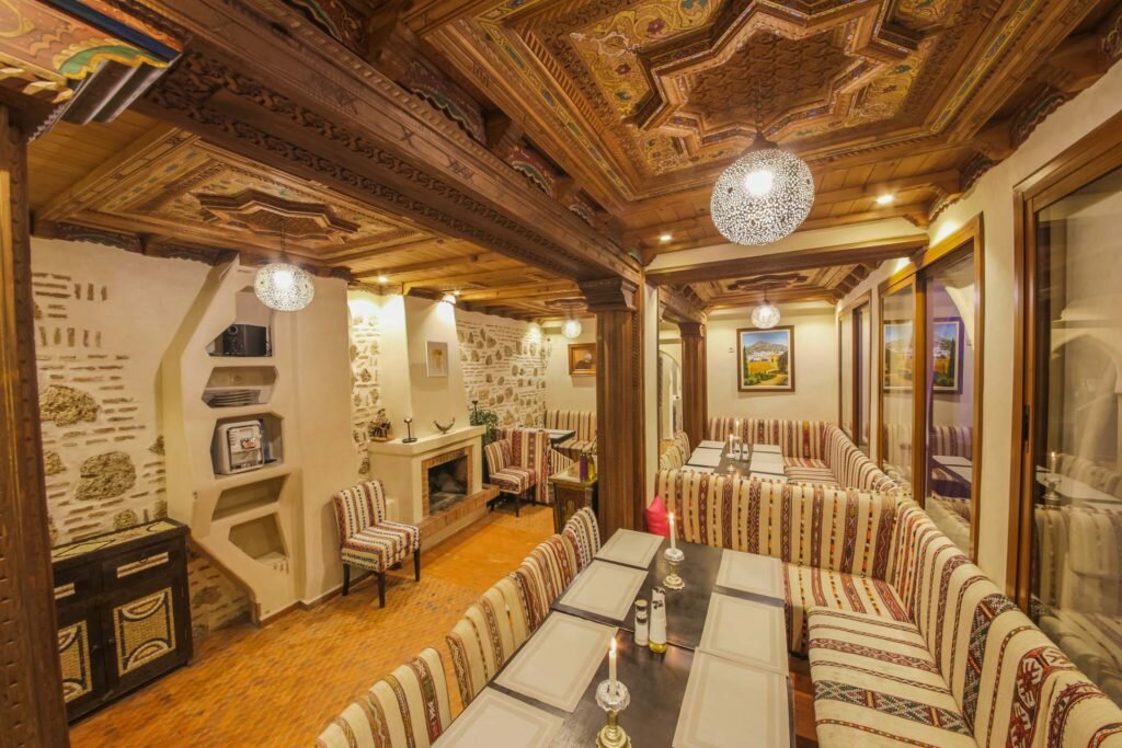 Rustic dining room with striped cushioned benches and ornate wooden ceiling. Soft lighting, framed paintings, candles, and warm ambiance.