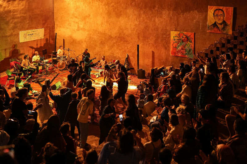 Concert in warm-lit hall, musicians on stage with guitars and drums in the moroccan sahara, crowd dancing and clapping, colorful artwork on walls. Energetic mood.