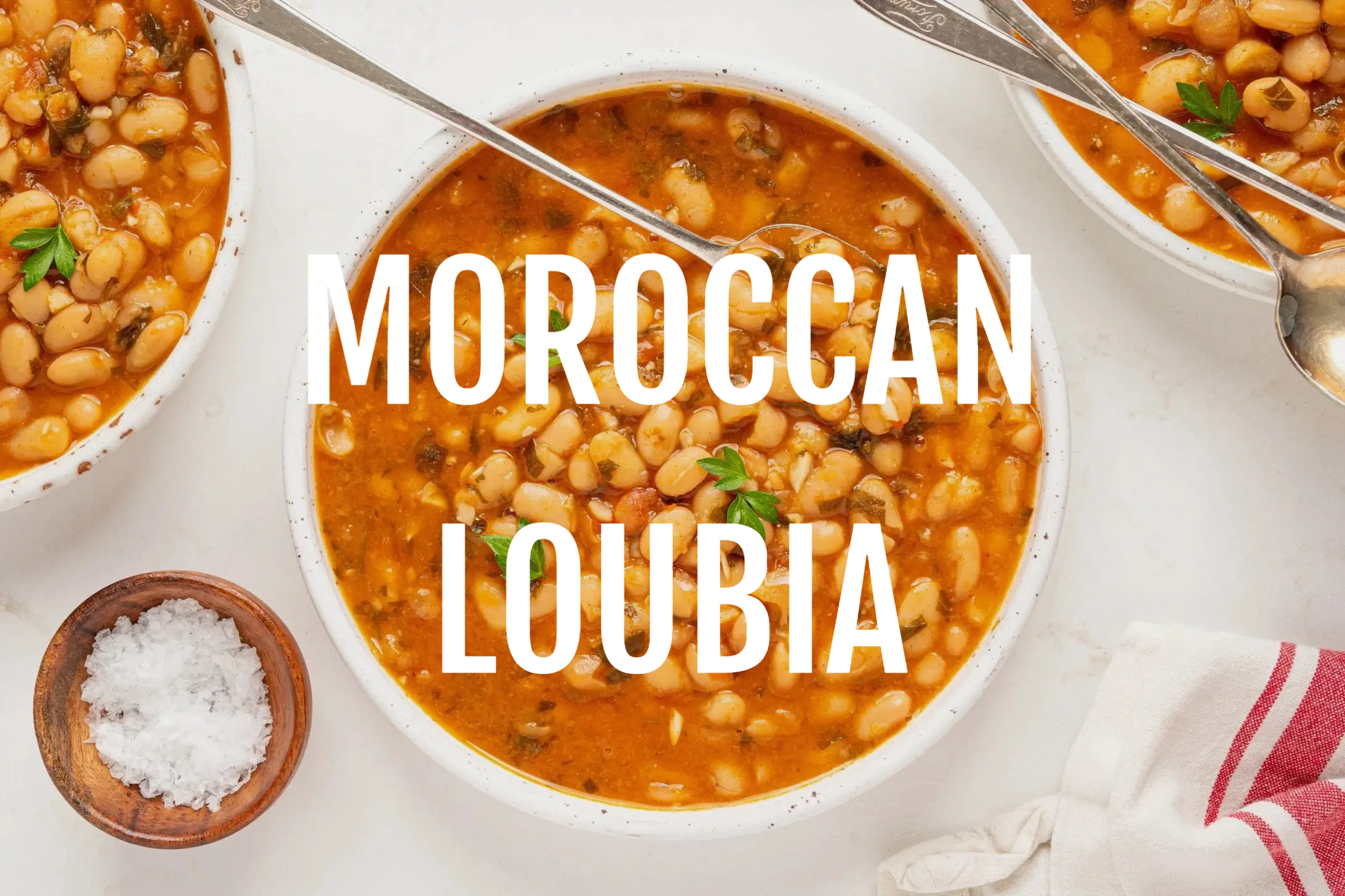 Bowl of Moroccan Loubia topped with parsley, surrounded by spoons, salt in a wooden bowl, and a red-striped cloth. Text ‘Moroccan Loubia’ overlays.