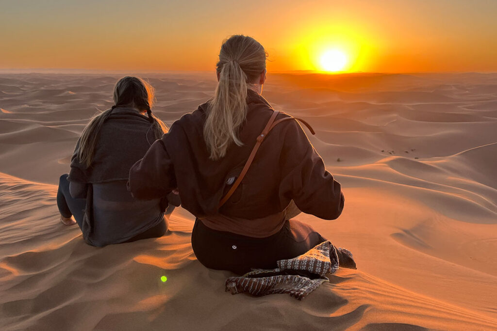 Two people sit on desert dunes at sunset in morocco, facing the sun. The sky is orange, casting warm light over the sandy landscape.