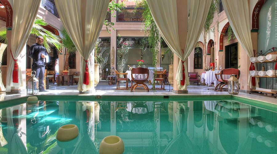 Elegant courtyard with a turquoise pool, white drapes, and tables set for dining. A person stands looking relaxed. Lush greenery and soft light.