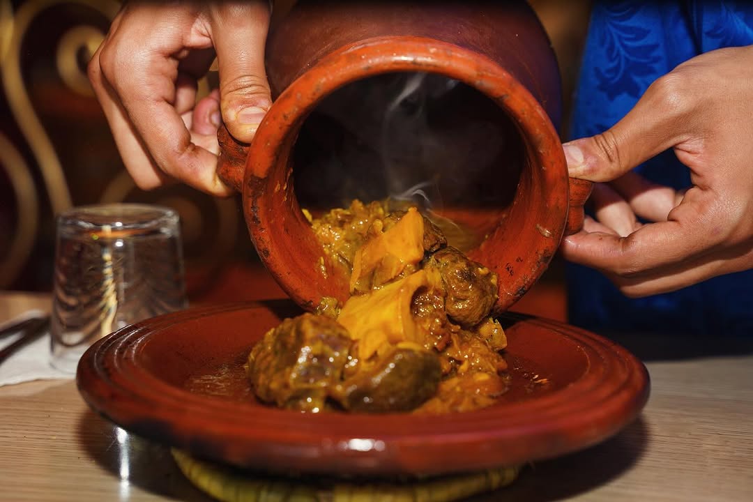 Hands pour steaming stew from a clay pot called tanjia onto a plate, with a glass in the background. Warm, rich colors suggest a cozy atmosphere in Morocco.