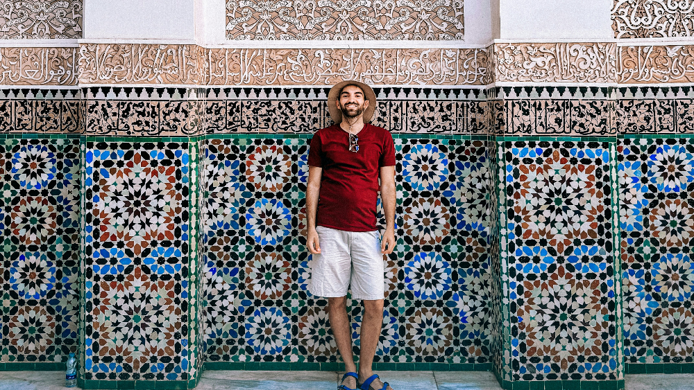 Man in a hat and red shirt stands smiling against a colorful, intricate tiled wall with ornate Arabic calligraphy above in morocco. Bright, relaxed setting.