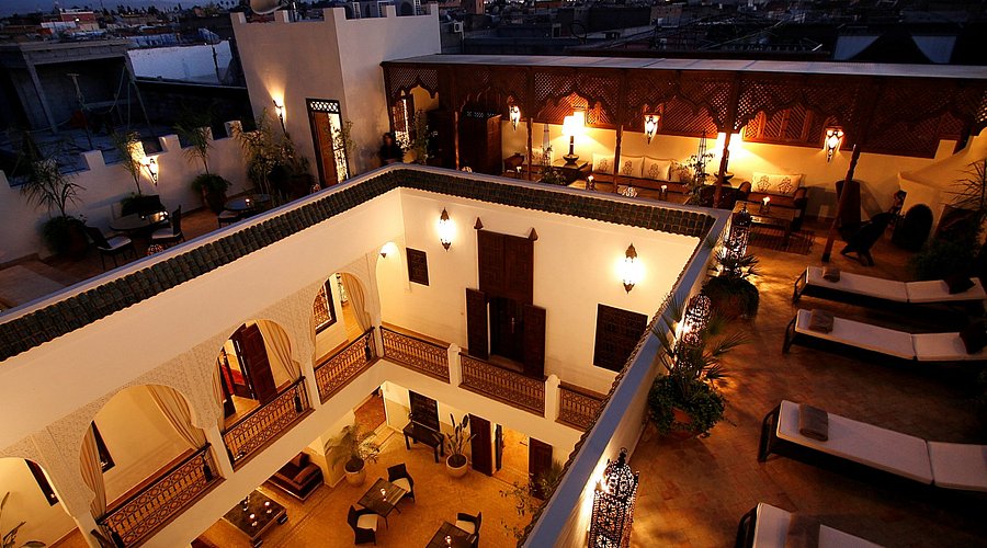 Lit courtyard at dusk with arched doorways, lanterns, and rooftop seating. Warm earthy tones create a cozy, inviting atmosphere.