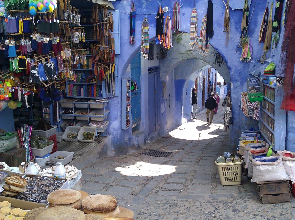 Narrow blue alley with vibrant market stalls, textiles hanging, people walking. Sunlit cobblestone street creates a lively, colorful scene.