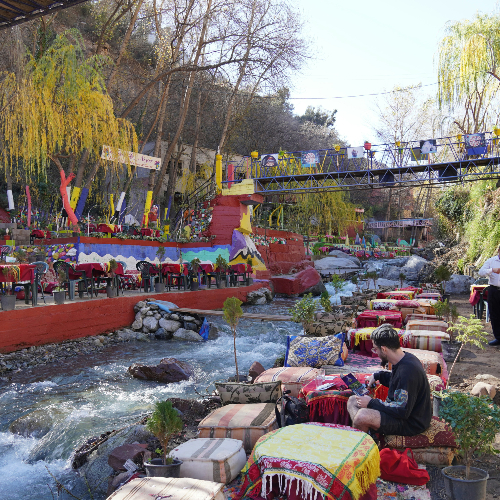 Colorful riverside café scene with a decorated bridge, vibrant tables, and a man seated near a flowing stream, surrounded by autumn trees.
