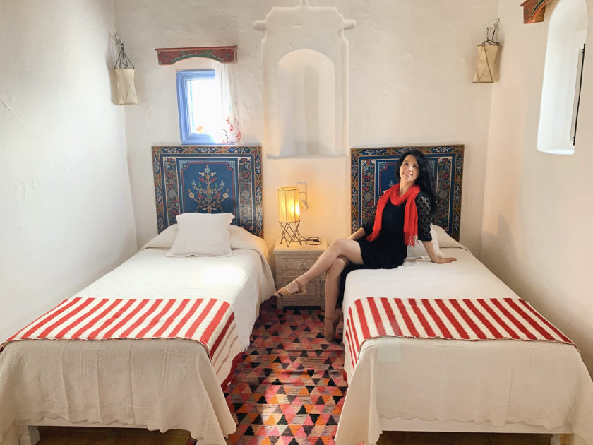 A woman in black with a red scarf sits on a bed in a cozy room with white walls, colorful headboards, and red-striped blankets.
