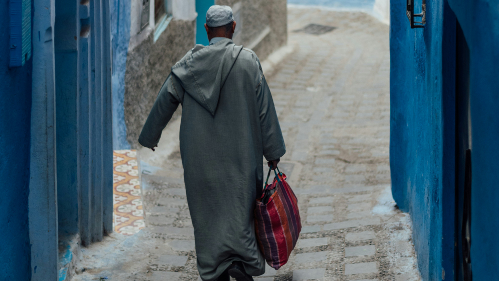 Moroccan man in gray jellaba and cap walking down a narrow, blue-painted alley, carrying a red-striped bag, creating a serene and calm atmosphere.