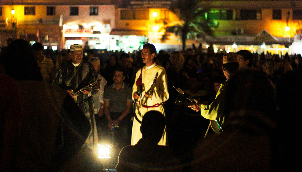 Traditional Moroccan Musicians performing with instruments outdoors at night, surrounded by a crowd in Marrakech, Morocco. Warm lights and buildings in the background create a lively mood.