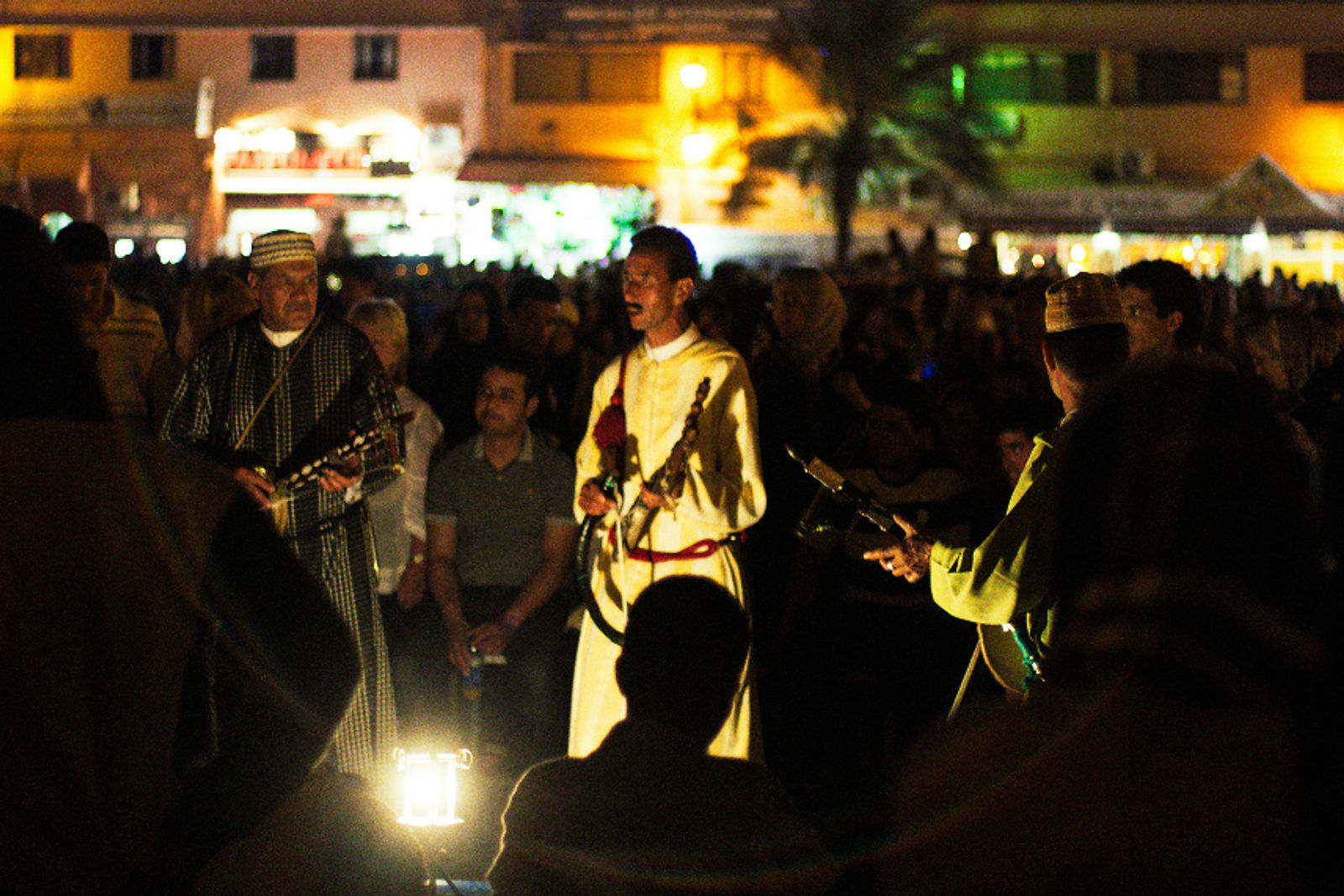 Traditional Moroccan Musicians performing with instruments outdoors at night, surrounded by a crowd in Marrakech, Morocco. Warm lights and buildings in the background create a lively mood.
