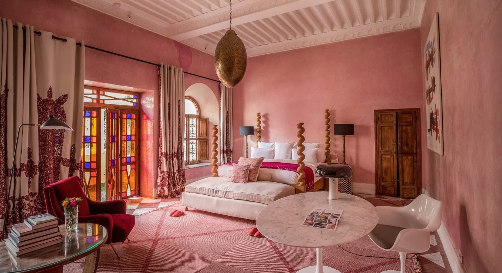 Pink bedroom with a plush bed, stained glass doors, and a marble table. A red chair holds flowers, and there’s warm, elegant decor.