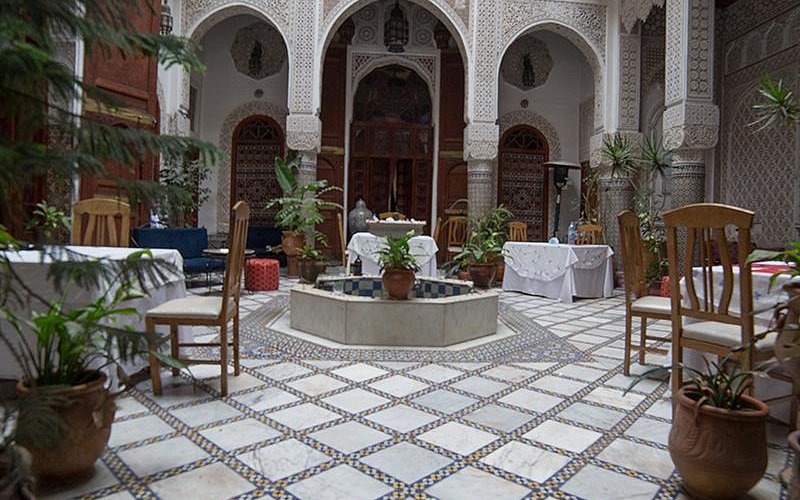 Ornate courtyard with tables, plants, and a central fountain. Arched doorways and intricate tiles add elegance. Serene, inviting atmosphere.
