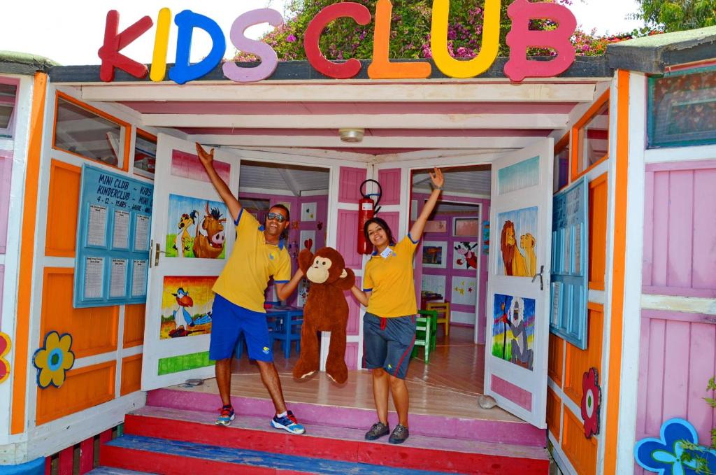 Two people in yellow shirts joyfully pose with a monkey toy at a colorful ‘KIDS CLUB’ entrance. Bright painted animals on doors.