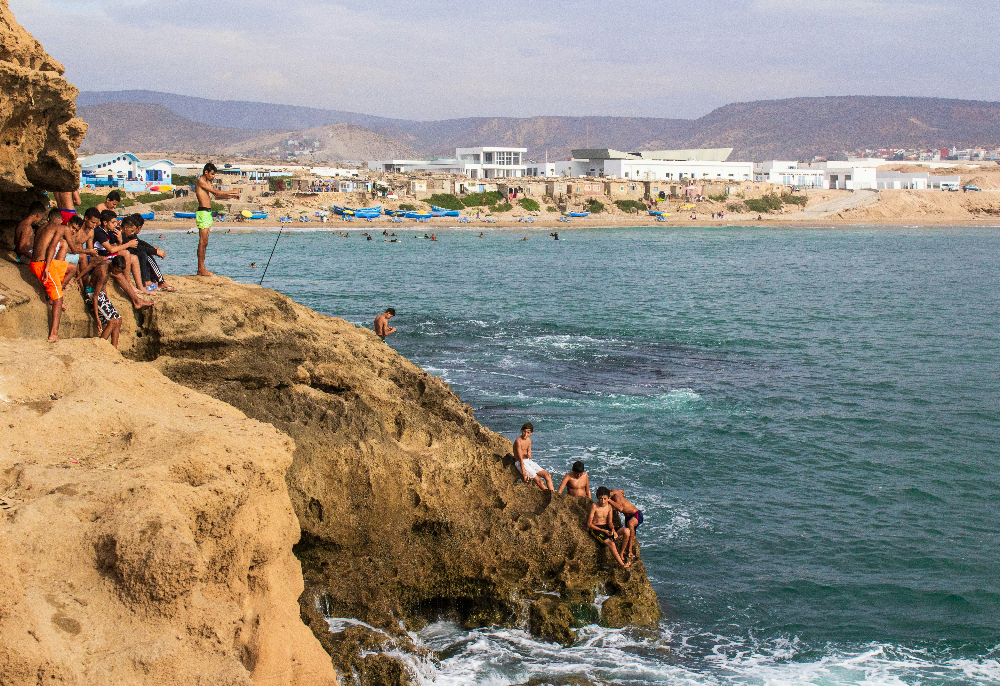 Teenagers on a rocky coast, some sitting and diving into the sea in Tamraght Morocoo. Background shows hills, buildings, and a sandy beach scene.