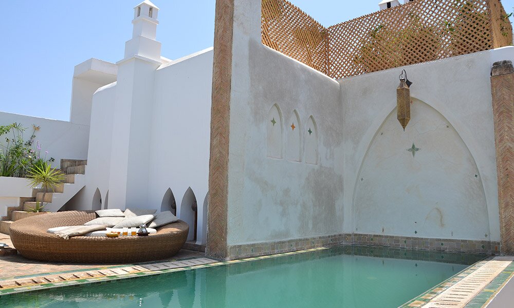 Circular wicker lounge with cushions by a turquoise pool, surrounded by white walls with arches. Bright, sunny, serene atmosphere.