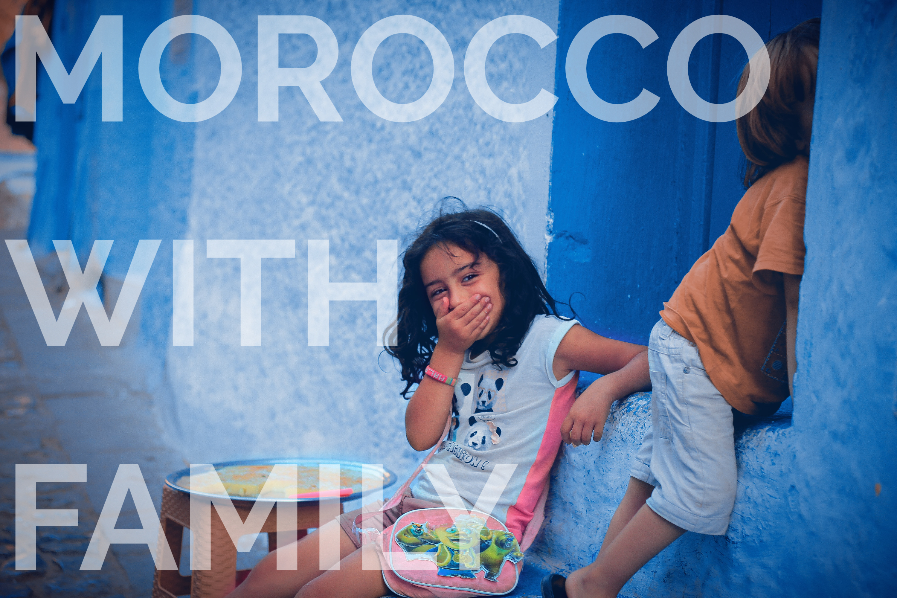 Girl smiles, covers mouth, sitting by blue wall with another child nearby. Text ‘Morocco with Family’ overlays. Playful mood.