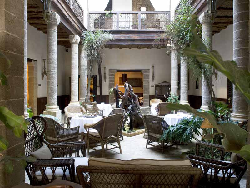 Elegant courtyard with wicker furniture and white tablecloths, surrounded by stone columns and lush plants, creating a serene ambiance.