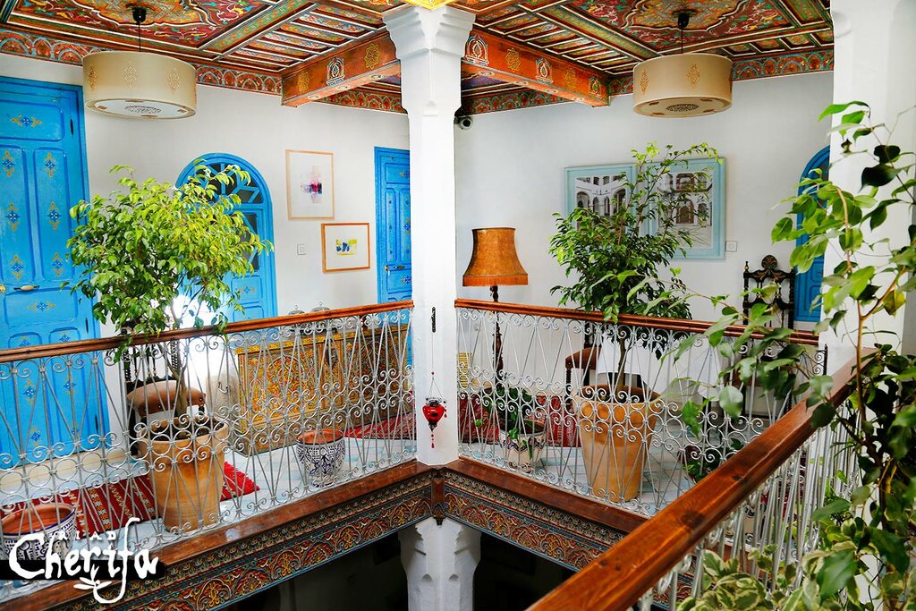 Colorful interior of a traditional house with ornate blue doors, vibrant plants, patterned ceiling, and a cozy atmosphere. Visible text: ‘Cherifa.’