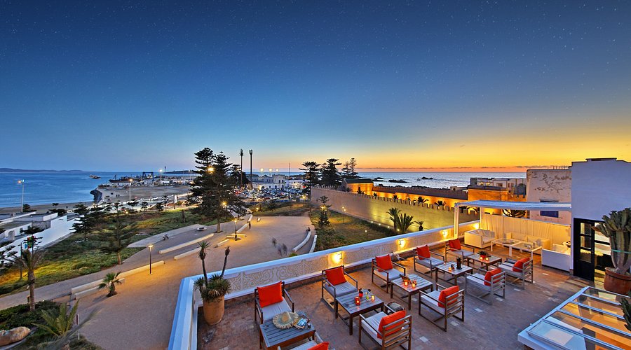 Rooftop terrace with orange chairs overlooks a coastal sunset. Lit pathways, ocean view, and starry sky create a serene ambiance.