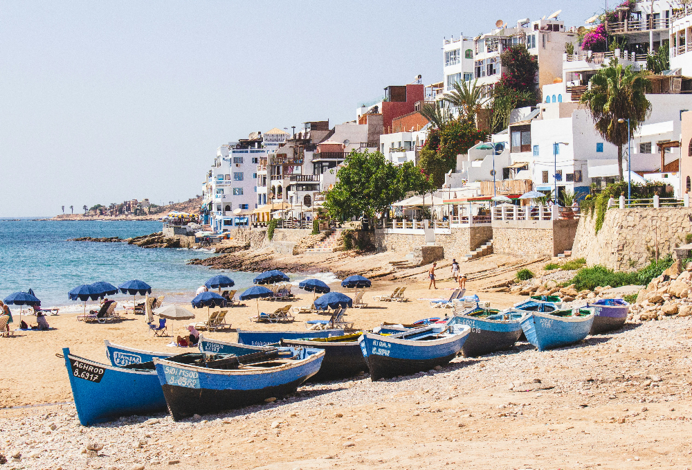 Boats on a sandy beach with blue umbrellas, people relaxing. White buildings line the shore with vibrant greenery under a clear sky in taghazou morocco