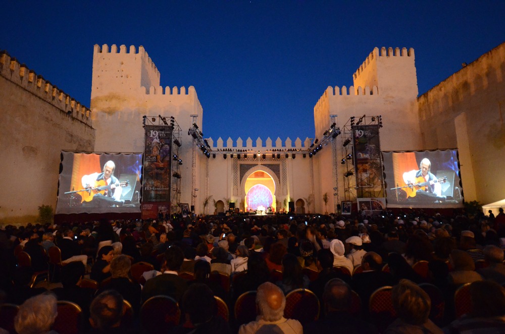 Outdoor concert at night in a courtyard in fes, morocco, with high walls; musician shown on large screens playing guitar; audience seated; vibrant blue sky.